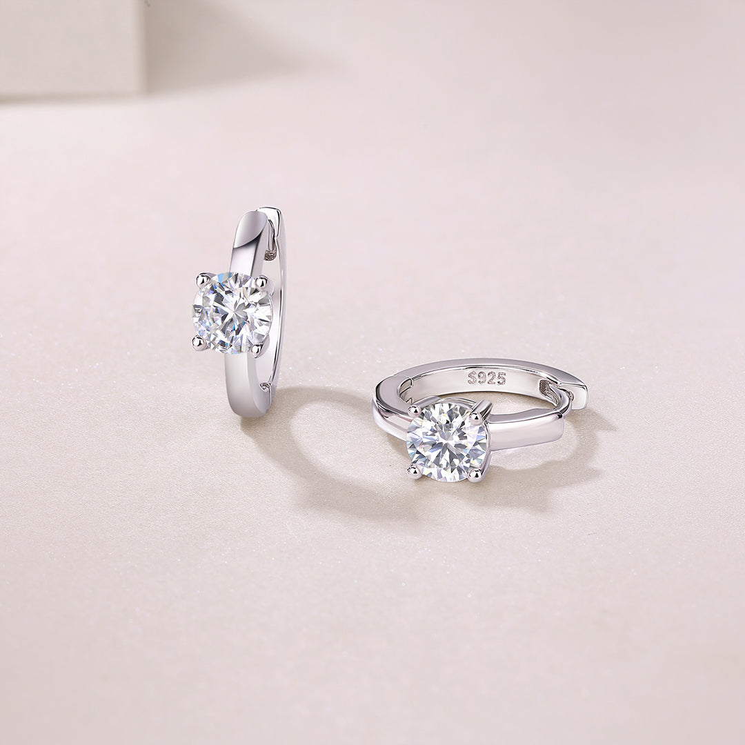 A pair of Éclat Solitaire Moissanite Huggie Earrings made with sterling silver and featuring round-cut moissanite stones, displayed against a neutral background.