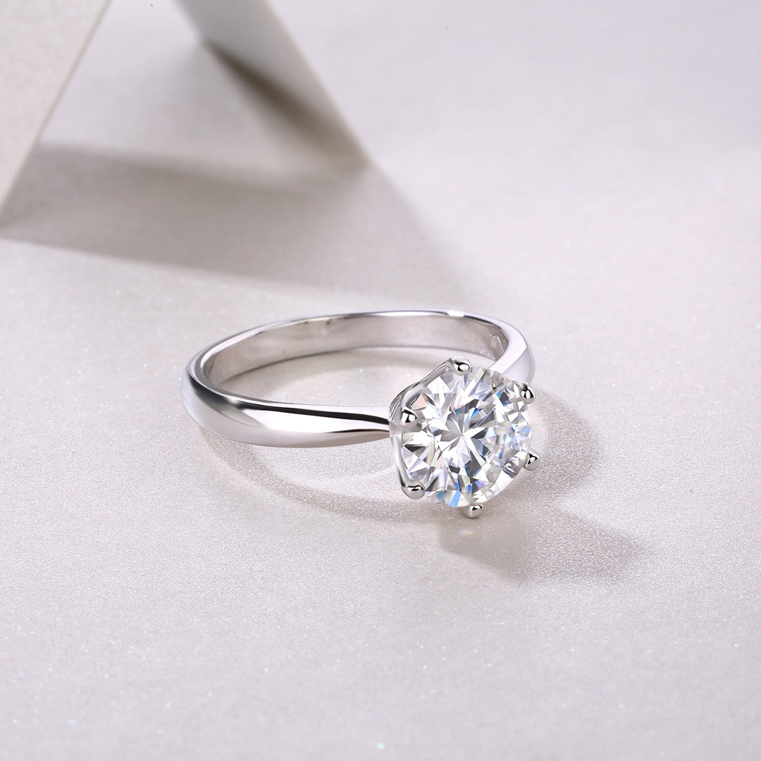 A silver solitaire ring with a round-cut moissanite stone in a six-prong setting, displayed on a white surface.