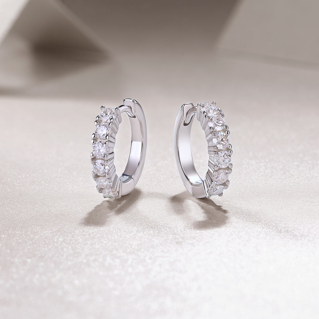 A pair of round-cut moissanite earrings with a huggie design, set in silver hoop settings, displayed against a white background.
