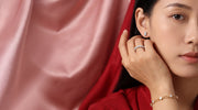 A close-up lifestyle shot of a woman wearing delicate gold jewelry, including a ring, bracelet, and earring, set against a backdrop of draped pink and red satin fabric.
